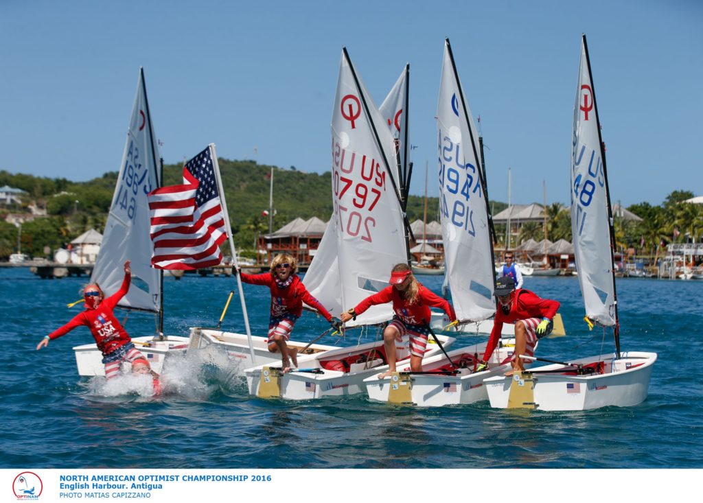 Four young sailors in red shirts celebrate on small sailboats, one waving a large American flag. The boats have numbered sails and are on blue water near a tropical shore with buildings and palm trees in the background.