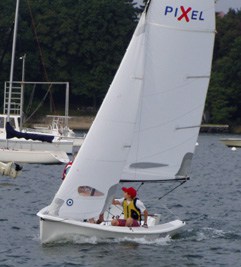 A person wearing a yellow life jacket and red cap is sailing a small white sailboat named PIXEL on a body of water, with other boats and trees visible in the background.