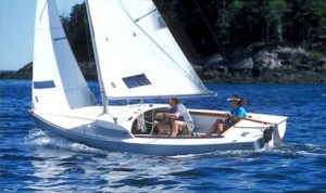 Two people are sailing a small white sailboat on calm blue water, with trees and rocky shore visible in the background. The sky is clear and sunny.