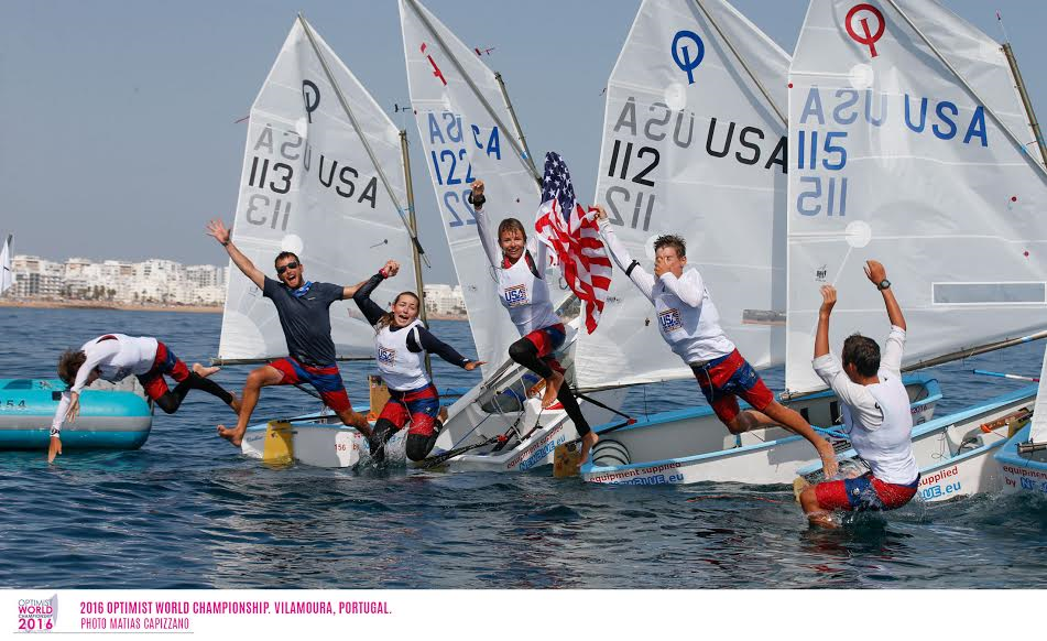 A group of young sailors in matching outfits celebrate on small sailboats, with one holding a U.S. flag. They cheer and gesture excitedly on the water, with a coastal city visible in the background.