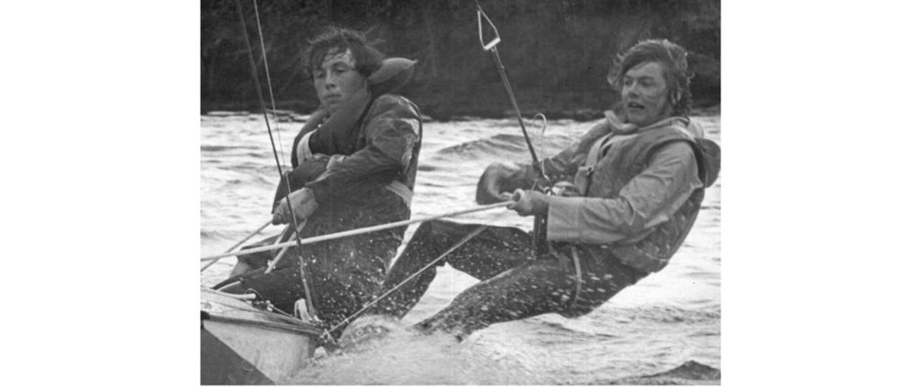 Two people wearing life jackets are sailing on a small boat in choppy water, leaning out over the side to balance. The image is in black and white.
