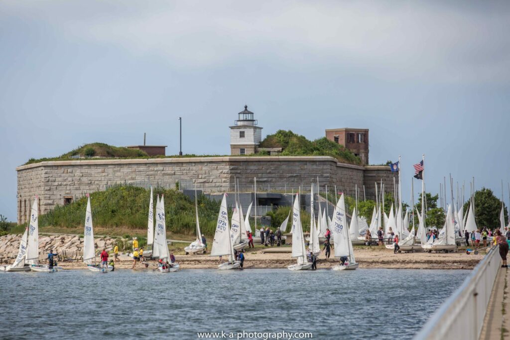 Sailboats gather near the shore in front of a historic stone fort with a lighthouse on top. People are standing by the boats, and flags are visible on the right side. The sky is partly cloudy.