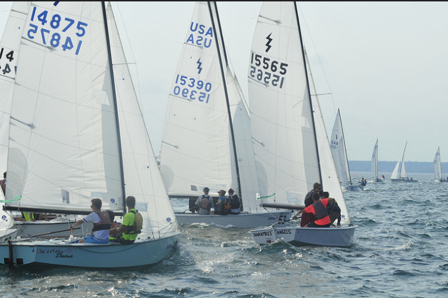 Several small sailboats with crews are racing on open water, with visible sail numbers and gentle waves. The background shows more boats and a distant shoreline under a slightly hazy sky.