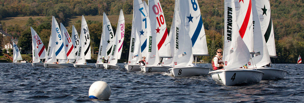 A group of small sailboats with numbered sails race across a lake, with sailors in life jackets. The scene is lively under a cloudy sky, with forested hills and houses in the background, and a buoy in the foreground.