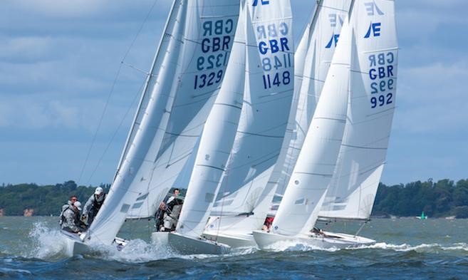 Three white sailboats with GBR sail numbers race closely on choppy water under a blue sky, with crews actively maneuvering. A green, tree-lined shore is visible in the background.