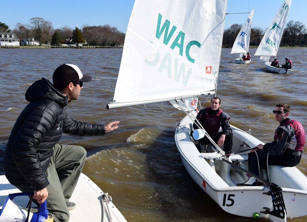 A man on a dock gestures toward two smiling sailors in a small sailboat marked 'WAC' and '15' on a windy, brown-water lake, with more sailboats and houses visible in the background.