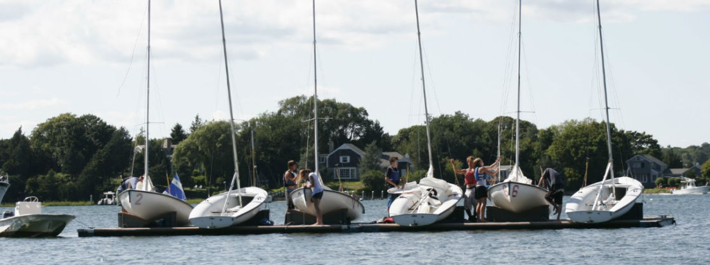 Six sailboats with people standing on a dock, preparing or adjusting the boats, with trees and houses in the background on a sunny day.