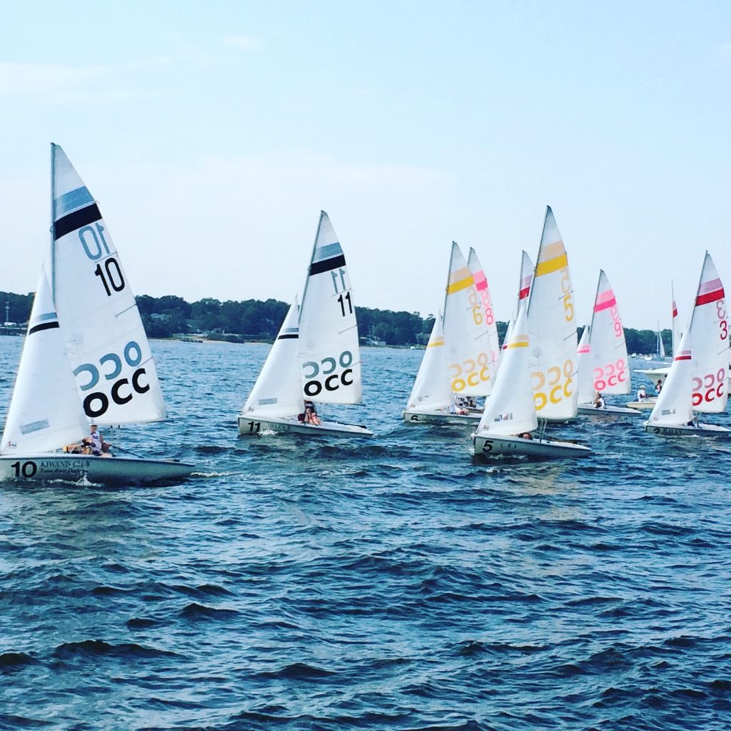 Several small sailboats with numbered sails race close together on a blue, wavy lake, with a tree-lined shore visible in the background under a mostly clear sky.