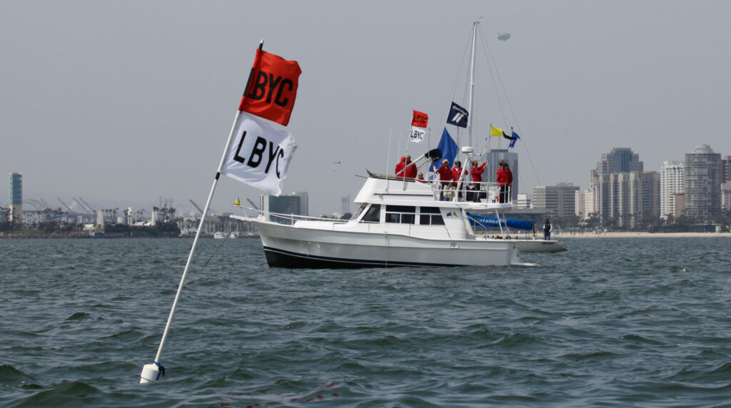 A white boat with people in red shirts and nautical flags on board floats on the water near a city shoreline. In the foreground, two buoy flags marked 'BYC' and 'LBYC' are visible in the water.