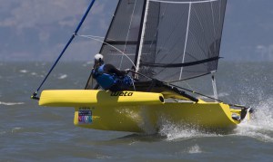 A person wearing a helmet sails a bright yellow, small trimaran with black sails at high speed over choppy water, with land visible in the distant background.