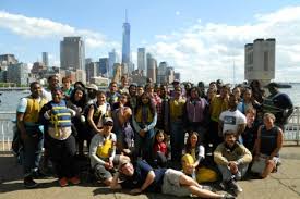 A large group of people poses together outdoors with tall city buildings and the One World Trade Center visible in the background on a sunny day. Some are wearing yellow vests; water is visible behind them.