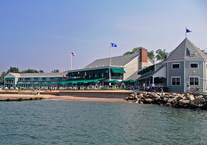 A waterfront building with green awnings and flags on the roof sits next to a sandy beach and rocky shoreline; people are visible on the terrace and near the water under a clear sky.
