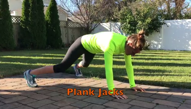 A person in a neon green long-sleeve shirt and black leggings performs plank jacks on a brick patio outdoors. The text 'Plank Jacks' appears at the bottom of the image. Trees and a fence are visible in the background.