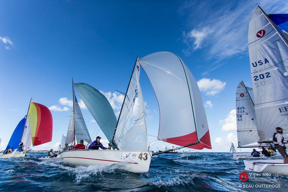 Several sailboats with colorful spinnakers race on open water under a blue sky. Sailors are actively maneuvering the boats, and waves create splashes around the hulls. Racing numbers and country codes are visible on sails.