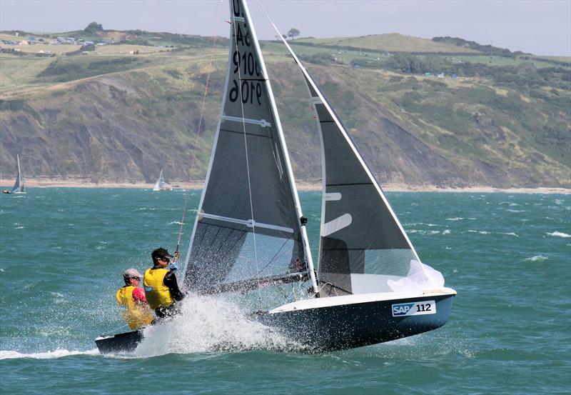 Two people in yellow life jackets sail a small racing boat, cutting through choppy blue water and creating a spray. Hills and cliffs are visible in the background under a partly cloudy sky.