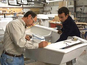 Two men work together in a workshop, measuring and marking a large white sink or bathtub. Tools and equipment are visible on the table and in the background.