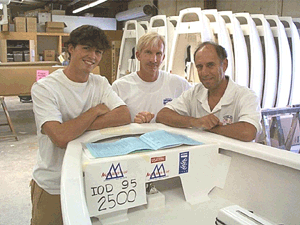 Three men wearing white shirts stand in a workshop, leaning on a small white boat with “IOD 95 2500” marked on it. Stacks of similar boats and workshop equipment are visible in the background.