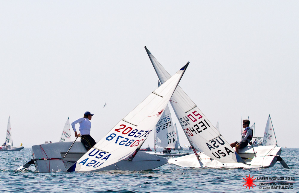 Two sailors in USA sailboats collide on the water during a race, causing both boats to tip and their sails to cross. Other sailboats are seen racing in the background under a clear sky.