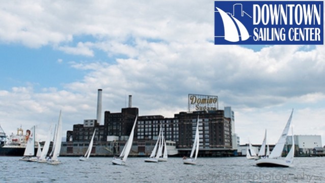 Several sailboats glide on the water in front of the Domino Sugar factory, with a partly cloudy sky above. The Downtown Sailing Center logo appears in the upper right corner.