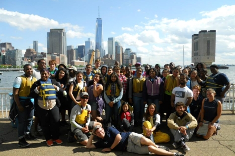 A large group of people poses together outdoors by a waterfront, with life jackets visible on some. Tall buildings and the One World Trade Center dominate the skyline in the background under a partly cloudy sky.