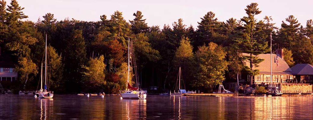 Sailboats float on a calm lake at sunset, with trees lining the shore and a large building with a dock and patio to the right. The scene is bathed in warm, golden light.