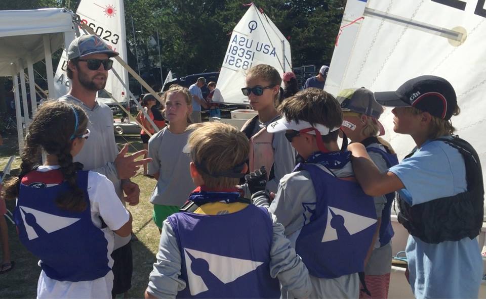 A coach talks to a group of young sailors wearing life jackets and sunglasses, standing together near small sailboats with sails marked with numbers at a regatta or sailing event.