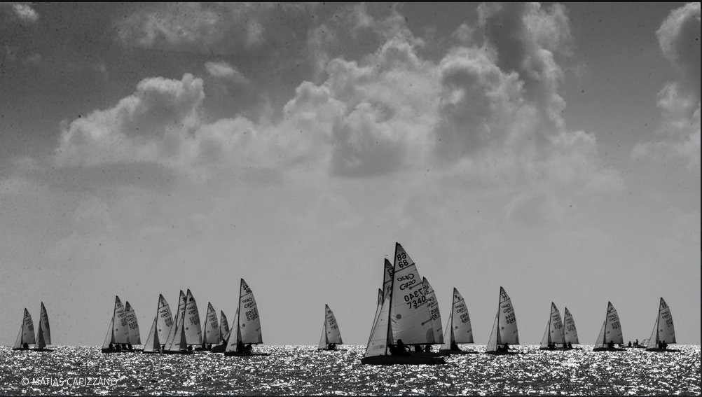 Black and white photo of numerous sailboats scattered across a shimmering sea, with dramatic clouds filling the bright sky above. The boats’ sails are illuminated by sunlight reflecting off the water.
