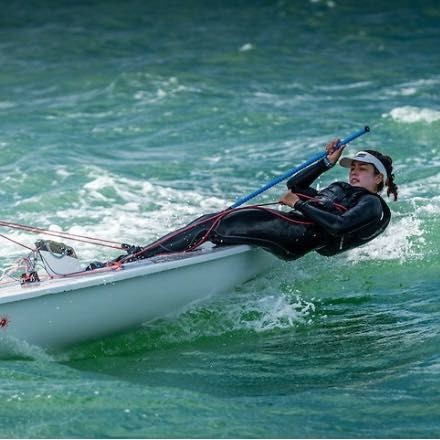 A person wearing a wetsuit and visor leans back over the edge of a small sailboat, holding a blue tiller, navigating through choppy green water.
