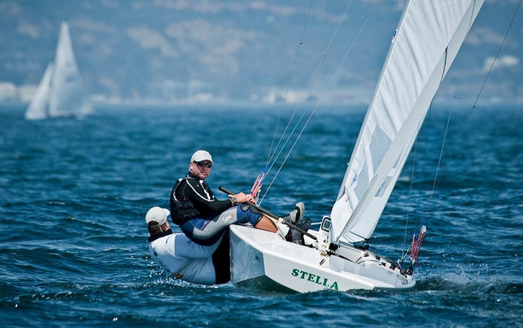 Two people sailing a small white boat named 'Stella' on blue water, with one person leaning out over the edge. Both wear wetsuits and sunglasses. Other sailboats and a blurred coastline are visible in the background.