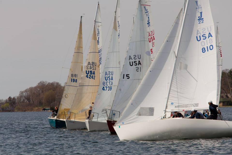 Four sailboats with white and tan sails, labeled with USA numbers, race closely together on a lake with trees and houses visible in the background. The sky is overcast and the water is calm.