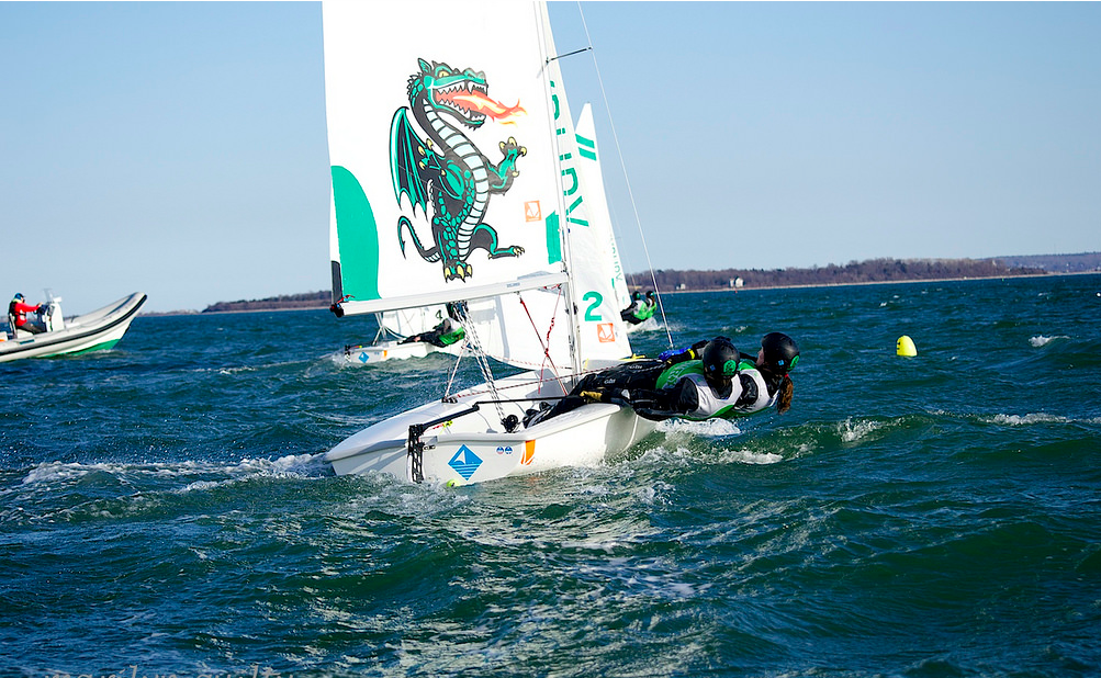 Two people sailing a small boat with a green dragon graphic on the sail, leaning out over the water on a windy day, with another boat and distant land visible in the background.
