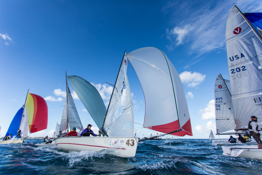 Several sailboats with colorful sails race across choppy blue water under a bright, partly cloudy sky. Sailors are actively steering and adjusting sails. The scene captures action and energy during a sailing competition.