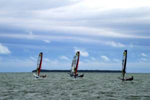 Three windsurfers glide across the water under a cloudy blue sky, with a distant landmass visible on the horizon.