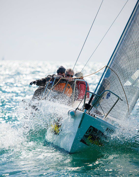 Three people sail a small boat through choppy waters, leaning to balance as ocean spray splashes high. The sunlight glistens on the water, and the sail is taut in the wind.