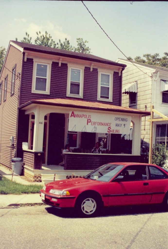 A red car is parked in front of a two-story brown house with a porch. A banner reads 'Annapolis Performance Sailing Opening May 9.' Trees and a neighboring house are visible in the background.