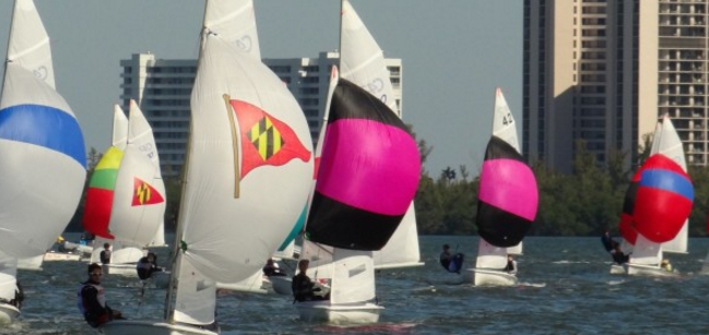 Sailboats with colorful sails race on the water near a city, with tall buildings and trees visible in the background. The sails are white with bright blue, pink, red, yellow, and green patterns.
