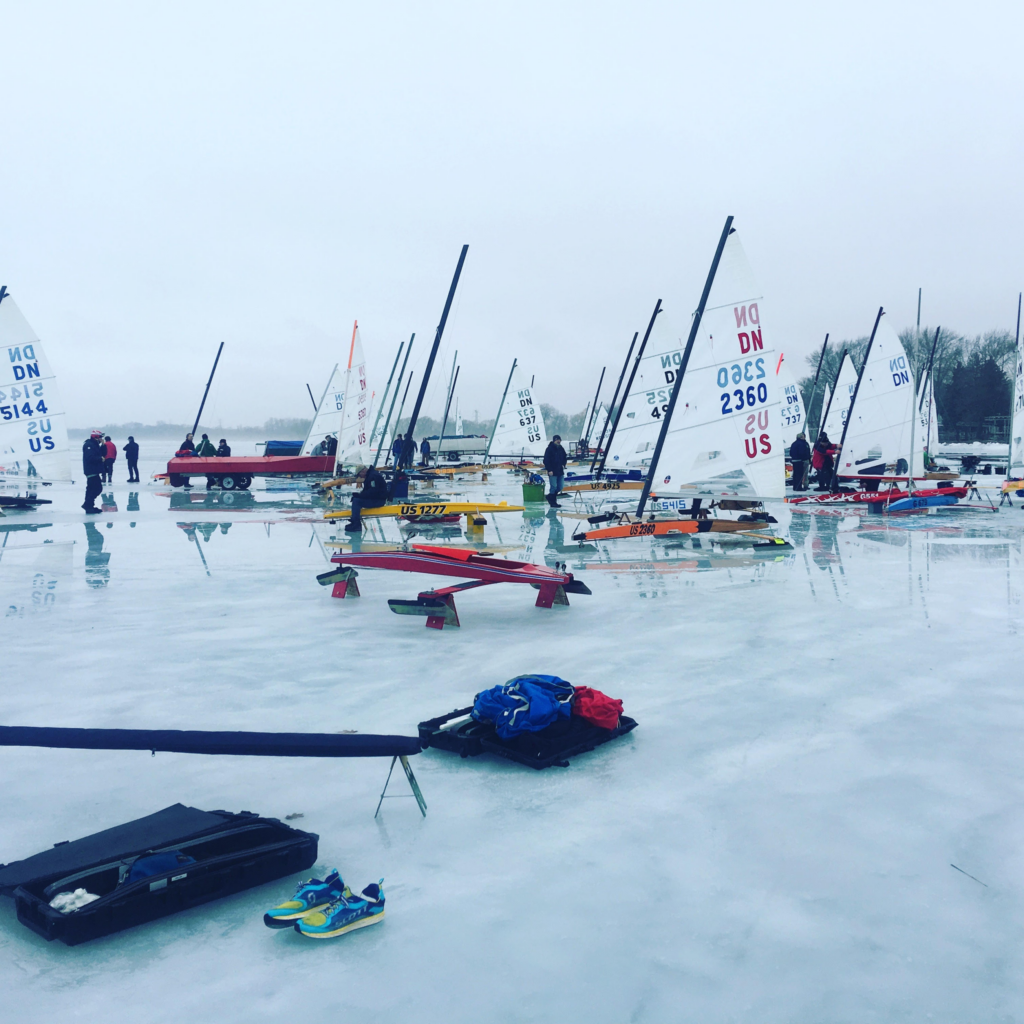 Iceboats with sails upright are lined up on a frozen lake, with people standing nearby. Gear and colorful shoes are scattered on the ice. The sky is overcast, creating a cold, wintry atmosphere.