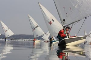 Several people sail small boats with white sails on calm water, close to the shore. The sky is overcast, and the water reflects the sails. The sailors wear jackets and life vests.