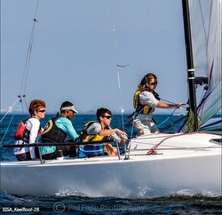 Four people wearing life jackets are sailing a white boat on open water. They are focused and working together, adjusting sails and steering under a clear blue sky.