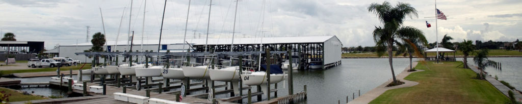 A marina with sailboats docked at piers, a covered boat storage building, palm trees, a grassy walkway, and American and Texas flags flying on a cloudy day.