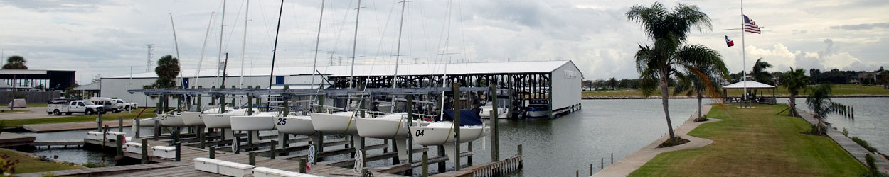 A marina with sailboats docked at piers, a covered boat storage building, palm trees, a grassy walkway, and American and Texas flags flying on a cloudy day.