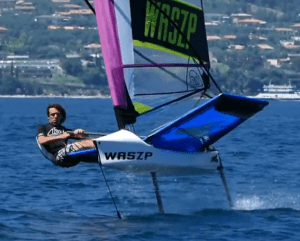 A person is sailing a hydrofoil boat labeled 'WASZP' over blue water. The boat is lifted above the water on foils, and the background shows a shoreline with houses and greenery.