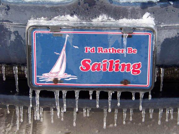 A license plate frame with the words “I’d rather be sailing” and an image of a sailboat, surrounded by icicles and frost, attached to a vehicle bumper.