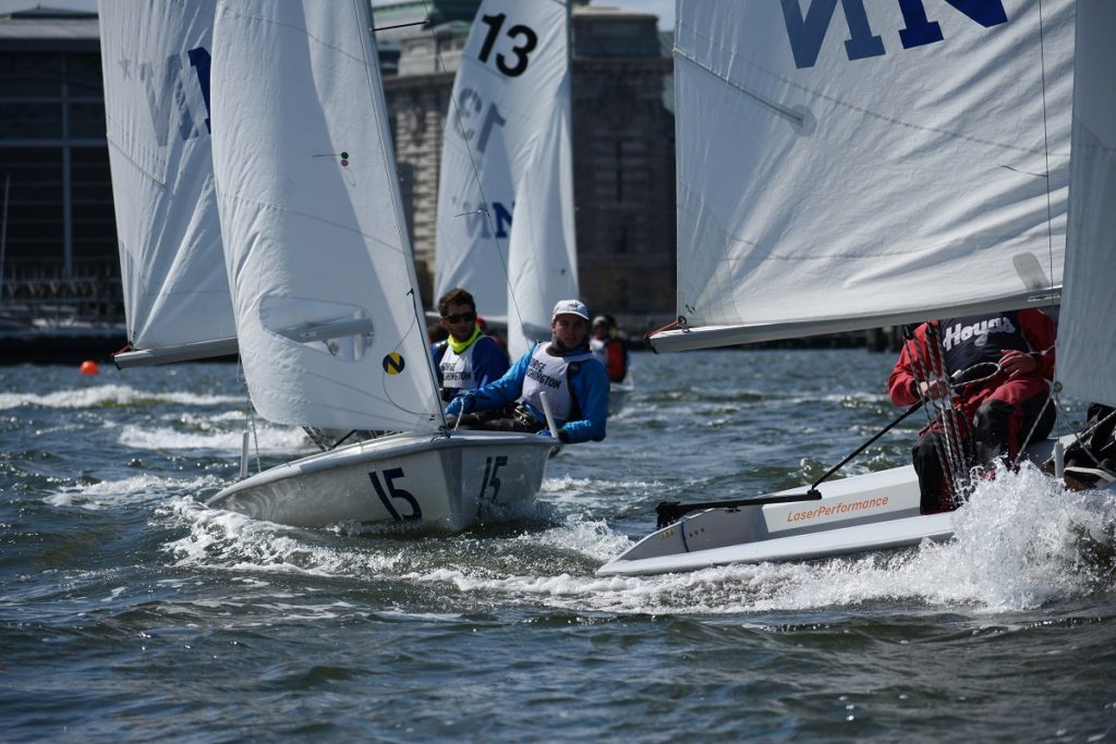 Three small sailboats with numbers compete closely on choppy water. Sailors wear life vests and helmets, focused as they navigate the race near a city waterfront with large buildings in the background.