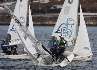 Three sailboats with 'CC CAMELS' logos compete closely on a body of water. Sailors in life jackets and gear manage the boats, with some leaning over the edge. Background shows a shore with buildings and trees.