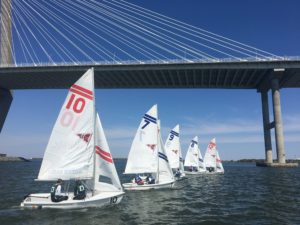Several small sailboats with numbered sails race on a river beneath a large cable-stayed bridge on a clear, sunny day. The water is calm, and the sky is bright blue.