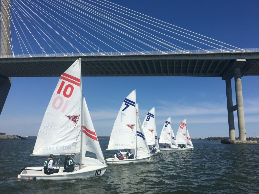 Several small sailboats with numbered sails race on a river beneath a large cable-stayed bridge on a clear, sunny day. The water is calm, and the sky is bright blue.