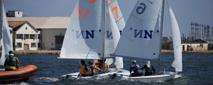 Two small sailboats with teams of sailors maneuver closely in a race near a large industrial building, while a person in a safety boat watches from the water. The sails display numbers and "N" logos.