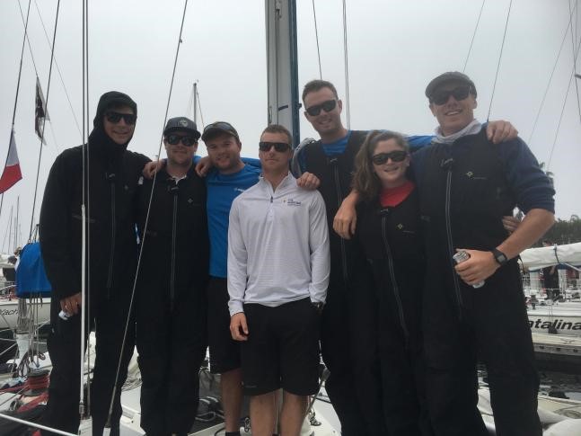 A group of seven people, dressed mostly in black sailing gear, stand together on a sailboat. Some are wearing sunglasses and hats. The background shows masts, rigging, and a cloudy sky at a marina.