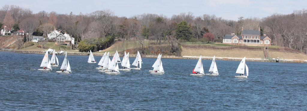 A group of small sailboats with white sails race across a wide river, with large houses and leafless trees visible on the opposite bank under a partly cloudy sky.
